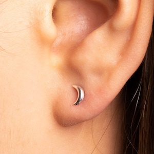 May include: A close-up of a person's ear with a small crescent moon earring. The earring is silver and is worn in the lobe of the ear.