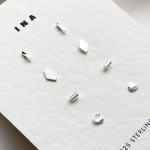 May include: A set of six silver earrings in various geometric shapes, including a diamond, a rectangle, a heart, and a circle. The earrings are displayed on a white card with the word "INA" and the text "925 Sterling Silver".