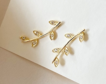 May include: A pair of gold-colored leaf-shaped earrings with small, clear gemstones. The earrings are displayed on a white card with the text "INA" and "925 STERLING SILVER". The background is a soft, off-white fabric.