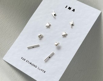 Könnte beinhalten: Ein Set aus 925er Sterlingsilber-Ohrringen mit verschiedenen Designs, darunter Herzen, Sterne und Blitze. Die Ohrringe werden auf einer weißen Karte mit dem Wort "INA" darauf präsentiert.