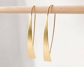 Könnte beinhalten: Ein Paar goldfarbene Ohrringe mit einem geschwungenen, flachen Design. Die Ohrringe hängen an einem Holzstift.
