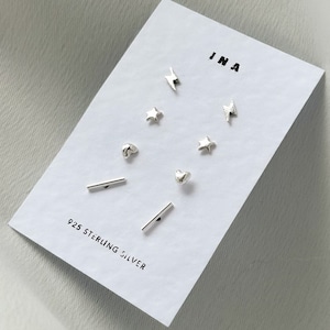 May include: A set of 925 sterling silver earrings featuring various designs, including hearts, stars, and lightning bolts. The earrings are presented on a white card with the word "INA" printed on it.