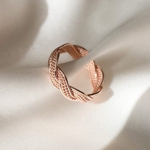 Puede incluir: Un anillo trenzado de oro rosa con un diseño delicado e intrincado. El anillo está hecho de metal y tiene un acabado liso y pulido.