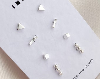 Könnte beinhalten: Ein Set mit sechs silbernen Ohrringen auf einer weißen Karte mit dem Text "INA" und "925 STERLING SILVER". Die Ohrringe beinhalten zwei Dreiecke, zwei Quadrate, zwei dünne Rechtecke und einen Satz mit drei kleinen silbernen Kugeln.