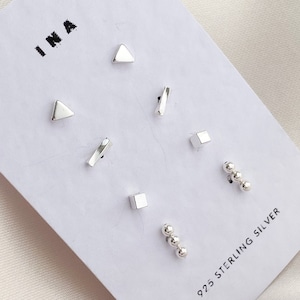 Peut inclure: Un ensemble de six boucles d'oreilles en argent sur une carte blanche avec le texte "INA" et "925 STERLING SILVER". Les boucles d'oreilles comprennent deux triangles, deux carrés, deux rectangles fins et un ensemble de trois petites boules d'argent.