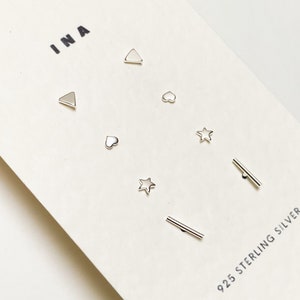 以下が含まれることがあります： ハート、星、三角形、線など、さまざまな形をした7つのシルバーイヤリングのセット。イヤリングは、「INA」と「925 STERLING SILVER」の文字が書かれた白いカードに飾られています。