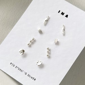 May include: A selection of sterling silver stud earrings in diverse geometric forms, such as bars, cubes, and spheres. The earrings are presented on a white card with the text "INA" and "925 STERLING SILVER".