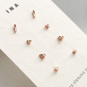May include: A set of eight gold-toned sterling silver earrings displayed on a white card with the text "INA" and "925 STERLING SILVER". The earrings feature a range of shapes, including squares, circles, and triangles.