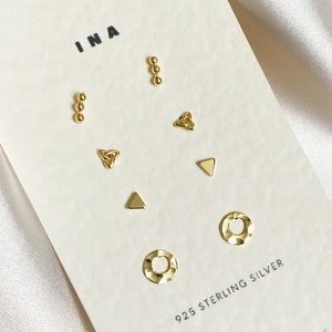 May include: A set of 925 sterling silver earrings with different designs, including a triangle, a circle, a heart, and a knot. The earrings are displayed on a white card with the word "INA" printed on it.