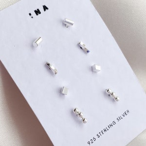 May include: A selection of minimalist sterling silver earrings on a white card. The earrings showcase diverse geometric forms, such as cylinders, squares, and curved bars, plus a set of three small ball studs. The card is labelled with "925 STERLING SILVER".