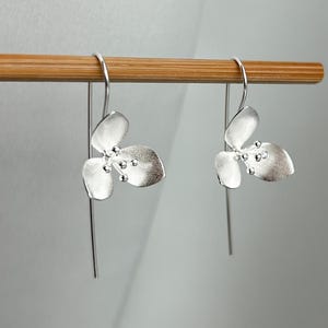 Könnte beinhalten: Ein Paar silberfarbene Ohrhänger. Jeder Ohrring hat ein florales Design mit drei Blütenblättern und kleinen, runden Details. Die Ohrringe hängen an einem dünnen, geraden Draht.