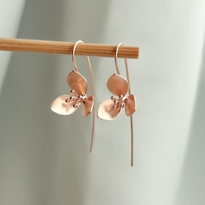 Puede incluir: Pendientes colgantes florales de oro rosa. Cada pendiente presenta tres elementos en forma de pétalo y pequeños detalles de cuentas. Los pendientes están suspendidos de un alambre curvo para la oreja y una caída larga y recta.