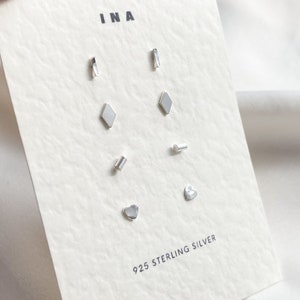 May include: A set of 925 sterling silver earrings with different shapes, including a heart, a diamond, a rectangle, and a line. The earrings are displayed on a white card with the word "INA" printed at the top.