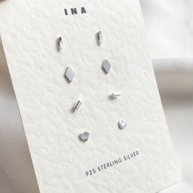 May include: A set of 925 sterling silver earrings with different shapes, including a heart, a diamond, a rectangle, and a line. The earrings are displayed on a white card with the word "INA" printed at the top.