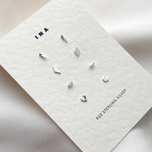 May include: A set of seven silver stud earrings in various shapes, including a heart, a diamond, and a rectangle. The earrings are displayed on a white card with the text "INA" and "925 STERLING SILVER".
