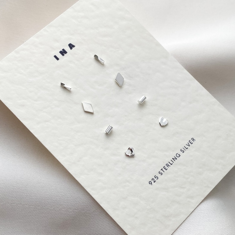 May include: A set of seven silver stud earrings in various shapes, including a heart, a diamond, and a rectangle. The earrings are displayed on a white card with the text "INA" and "925 STERLING SILVER".