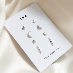 May include: A set of six silver earrings on a white card. The earrings are in the shapes of a heart, a crescent moon, a star, and a bar. The card has the text "INA" and "925 STERLING SILVER" printed on it.