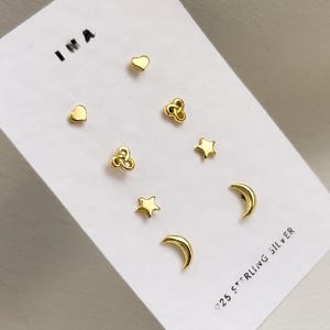 Puede incluir: Un conjunto de pendientes dorados con forma de corazón, trébol, estrella y media luna. Los pendientes se muestran en una tarjeta blanca con el texto "925 STERLING SILVER".