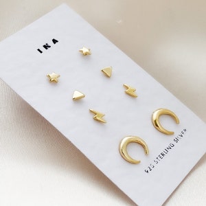 May include: A set of six gold-toned sterling silver earrings. The earrings feature a variety of shapes, including stars, triangles, lightning bolts, and crescent moons. The earrings are packaged on a white card with the brand name "INA" and the text "925 Sterling Silver".