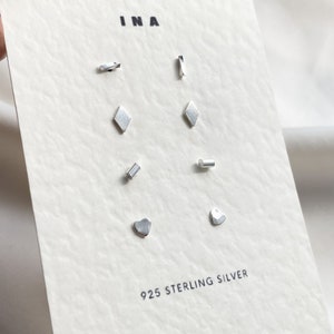 May include: A set of eight silver earrings with different geometric shapes, including a diamond, a heart, a rectangle, and a curved line. The earrings are displayed on a white card with the text "INA" and "925 STERLING SILVER".