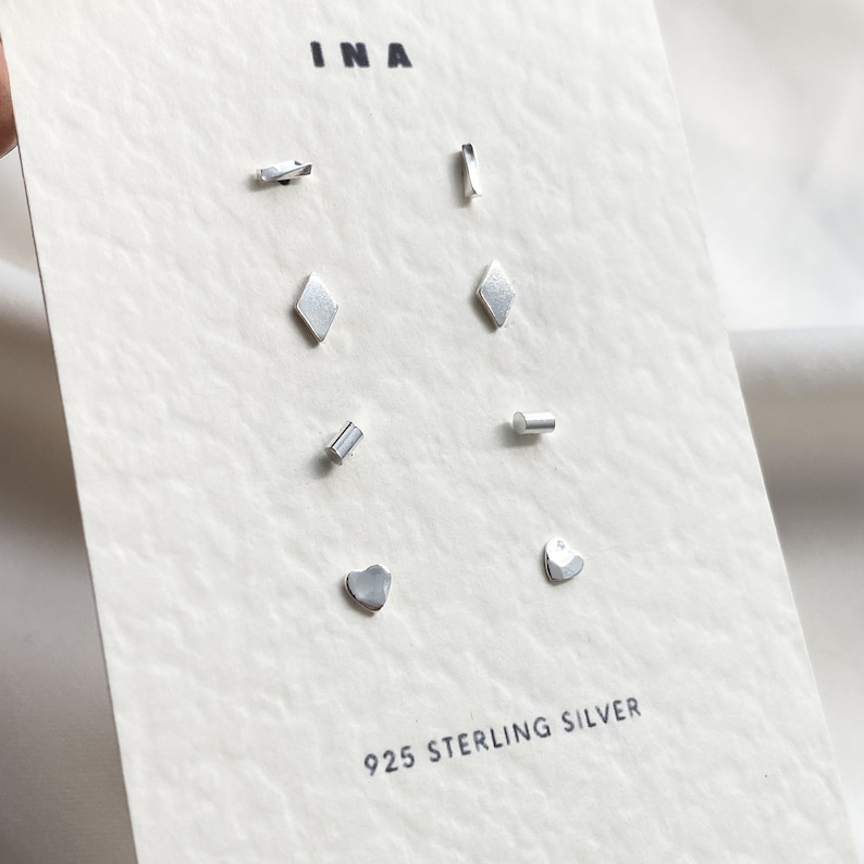 May include: A set of eight silver earrings with different geometric shapes, including a diamond, a heart, a rectangle, and a curved line. The earrings are displayed on a white card with the text "INA" and "925 STERLING SILVER".