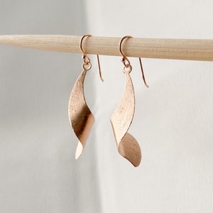 May include: Rose gold earrings with a twisted, leaf-shaped design. The earrings are hanging from a wooden surface.