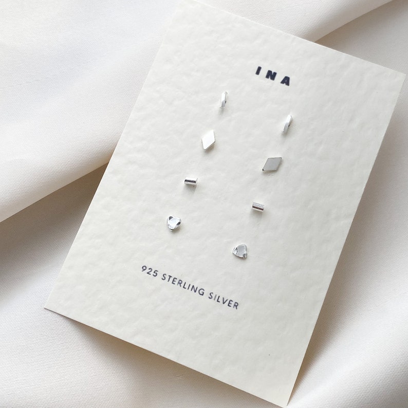 May include: A set of 925 sterling silver earrings in various shapes, including a heart, a diamond, and a rectangle. The earrings are displayed on a white card with the brand name "INA" printed on it.