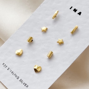 May include: A set of six gold-toned sterling silver earrings with different shapes, including a star, a heart, and a rectangle. The earrings are displayed on a white card with the text "INA" and "925 STERLING SILVER".