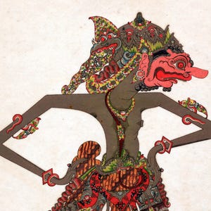 Op de afbeelding: Een kleurrijke, traditionele Indonesische Wayang Kulit-pop. De pop heeft een rood gezicht, een uitgebreide hoofdtooi en gedetailleerde kleding met rode, groene en gele accenten. De armen van de pop zijn uitgestrekt, tegen een witte achtergrond.