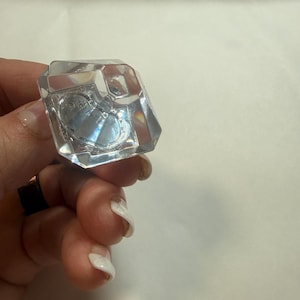 May include: A clear, faceted, diamond-shaped knob with a geometric design. The object is transparent and features multiple angled surfaces that reflect light, creating a prismatic effect. It has a flat top and a tapered base for mounting.