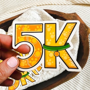 May include: A yellow and orange sticker with the number 5K printed on it. The number 5 is decorated with paw prints and the letter K has a green collar with a small orange bell hanging from it.