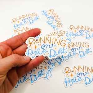 May include: A close-up of a hand holding a sticker that says "RUNNING on PIXIE DUST" in gold and blue lettering with stars. The sticker is surrounded by other stickers with the same design.