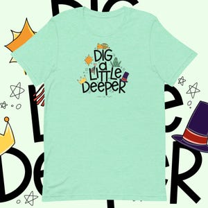 Op de afbeelding: Een lichtgroen T-shirt met de tekst "Dig a Little Deeper" in zwarte letters. De tekst wordt omringd door kleine illustraties van een kroon, een hart, een ster, een kikker, een trompet en een hoge hoed.