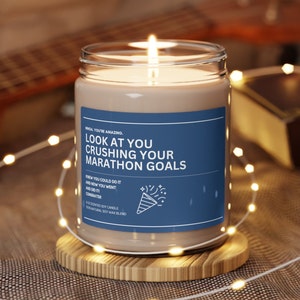 Puede incluir: Una vela de soja marrón con una etiqueta azul que dice "WHOA, YOU'RE AMAZING. LOOK AT YOU CRUSHING YOUR MARATHON GOALS. KNEW YOU COULD DO IT AND NOW YOU WENT AND DID IT! CONGRATS!"  9 oz scented soy candle. 100% natural soy wax blend.