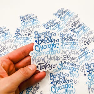 May include: A collection of blue and white stickers with the names of cities written in a stylized font. The cities include Boston, Berlin, Chicago, London, New York, and Tokyo.