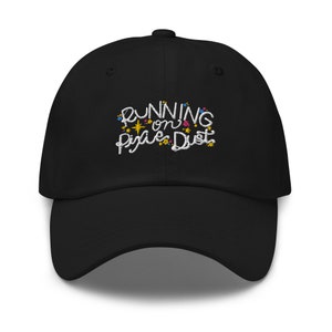 Op de afbeelding: Zwarte baseballpet met witte geborduurde tekst die "Running on Pixie Dust" zegt met kleurrijke glitteraccenten.