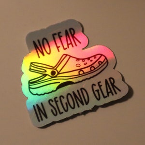 May include: Holographic sticker featuring a black outline of a Croc shoe with the text "NO FEAR IN SECOND GEAR". The sticker has a rainbow sheen, reflecting various colors. The design is on a white, cloud-shaped background.