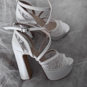 May include: A pair of white platform heels with lace detailing. The heels are open-toe and have a strap around the ankle.
