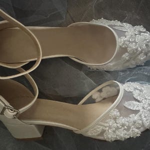 May include: A pair of white bridal shoes with delicate lace detailing and a low block heel.