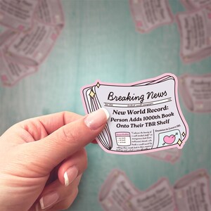 May include: A pink and white sticker with a newspaper design. The headline reads "Breaking News: New World Record: Person Adds 1000th Book Onto Their TBR Shelf".