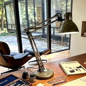 May include: An olive green metal desk lamp with an adjustable articulated arm and a dome-shaped shade. The lamp features a pull chain switch and sits on a circular base. It is positioned on a wooden desk next to a notebook labeled LE CORBUSIER.