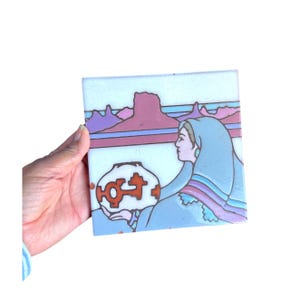 May include: A square ceramic tile featuring a graphic illustration of a person in a blue head covering holding a white vessel with orange geometric symbols. The background depicts a desert landscape with purple mesas under a light blue sky.