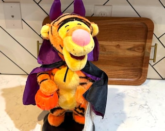 May include: A plush Tigger toy dressed as a vampire, featuring orange and black stripes, a pink nose, and purple devil horns. The character wears a black cape with a purple lining and holds a small orange plastic jack-o'-lantern bucket.