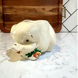 May include: A cream-colored ceramic piggy bank featuring a glossy finish with visible fine crackle patterns. The pig is depicted in a seated position with a painted green leaf and orange carrot detail near its snout and dark painted eyes.
