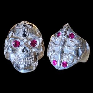 Può includere: Due anelli d'argento. Un anello è un teschio con occhi di gemme rosse e una croce sulla fronte. L'altro anello ha un design a scudo con gemme rosse. Entrambi gli anelli sono su sfondo nero.