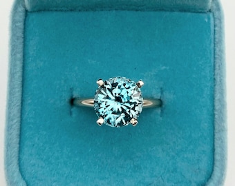 May include: A round, light blue gemstone ring set in a gold band, displayed in a teal velvet ring box. The gemstone is faceted, reflecting light. The ring is a classic solitaire design.