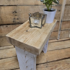 Peut inclure: Un petit banc en bois rustique avec un dessus en bois naturel et des pieds blancs. Un photophore en forme d'étoile et une petite plante en pot sont posés sur le banc. Le banc est de style ferme et est en bois récupéré.