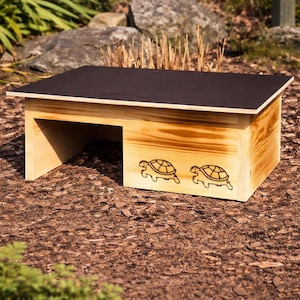 Peut inclure: Un habitat en bois pour reptiles avec un dessus marron foncé et une base marron clair. L'avant de l'habitat présente deux motifs de tortues gravés. L'habitat est conçu pour les petits reptiles et est placé à l'extérieur.