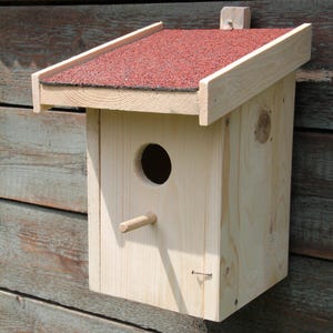 Könnte beinhalten: Ein Vogelhaus aus Holz mit einem roten Schindeldach, das an einer verwitterten Holzwand befestigt ist. Das Vogelhaus hat ein rundes Eingangsloch und einen kleinen Sitzplatz.
