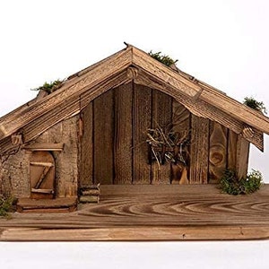 Può includere: Un presepe in legno con un tetto spiovente marrone e una piccola porta. La scena è posizionata su una base in legno con una superficie testurizzata.